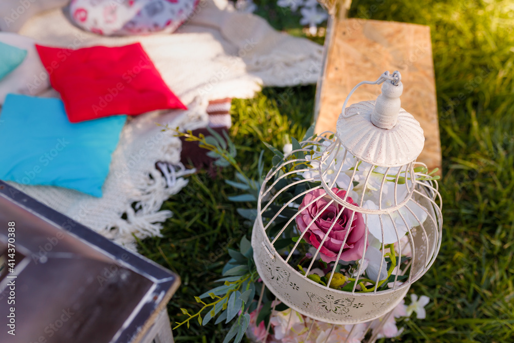 Fototapeta premium Soft focused shot of decorative bird cage filled with flowers, blurry cushions on plaid. Outdoors picnic party, birthday, proposal, anniversary.