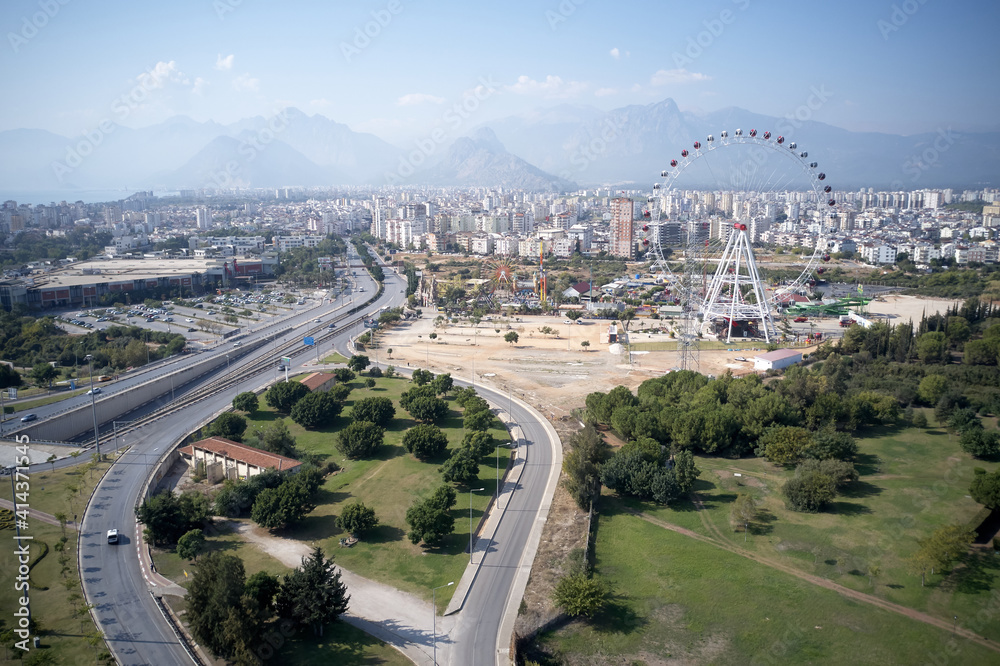 Urban landscape of Antalya town. Panoramic view of city buildings, road, Ferris wheel and mountains in the background.