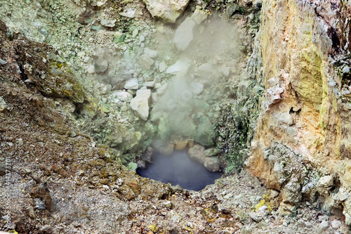 A crater filled with water with rising steam, around a wall with green and yellow stones