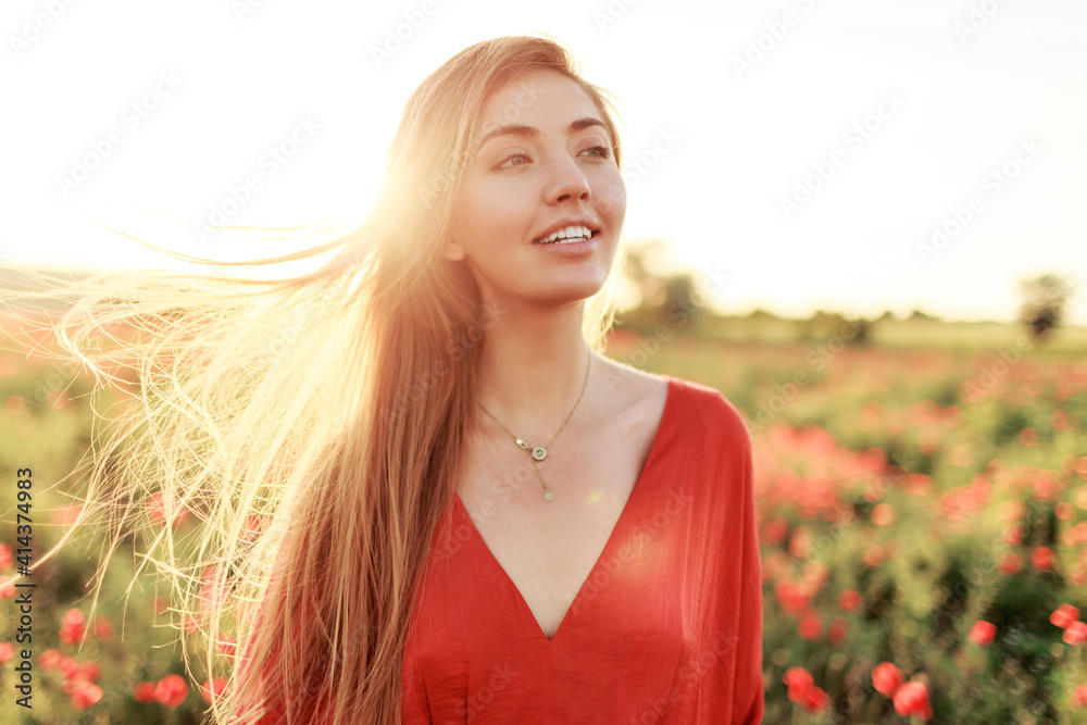 Gentle blonde long-haired woman with perfect smile posing on poppy field in warm summer sunset. - obrazy, fototapety, plakaty 
