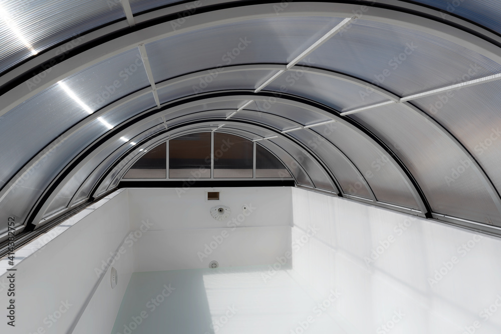 Fototapeta premium An empty fiberglass swimming pool with a sliding polycarbonate cover on a sunny spring day. A pool is white-colored, while the pavilion is half-transparent with blue toning.