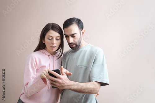 Two male and female adults being surprised shocked looking at the phone screen. Online shopping, having fun. Wearing casual pastel pink hoodie. Empty background, copy space for text, studio shot, clos