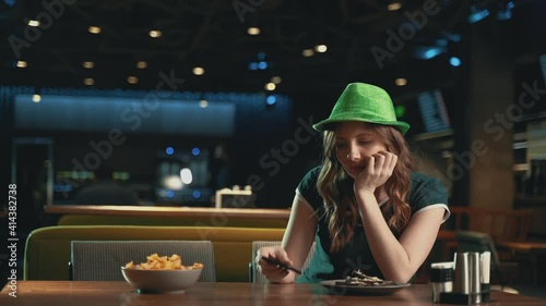 Pretty young woman waiting for someone, worried, looking at the phone and wearing an Irish green hat. Celebrating Saint Patrick's Day in a pub. 
