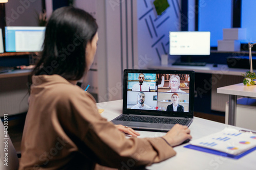 Entrepreneur talking in the course of online meeting with colleagues doing overtime. Woman working on finance during a video conference with coworkers at night hours in the office.