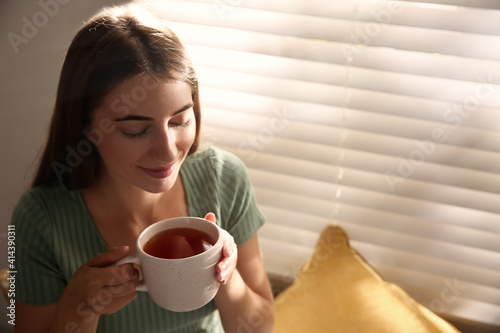 Beautiful young woman with cup of tea near window at home