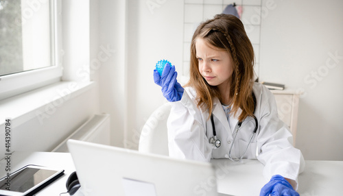 pretty blonde schoolgirl girl plays doctor and has white coat on and blue protective gloves and watches a blue ball  a model of a virus