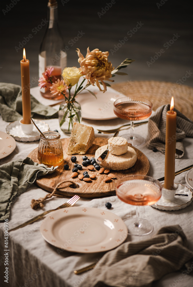 Romantic table setting, home lockdown party concept. Rose wine in bottle and glasses, cheese board, nuts, jam, dinnerware, cutlery, candles on light linen tablecloth, selective focus