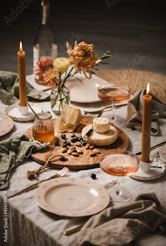 Romantic table setting  home lockdown party concept. Rose wine in bottle and glasses  cheese board  nuts  jam  dinnerware  cutlery  candles on light linen tablecloth  selective focus