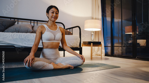 Young Asia lady in sportswear doing yoga exercise working out in living room at home at night. Sport and recreation activity, social distancing, quarantine for corona virus prevention concept.