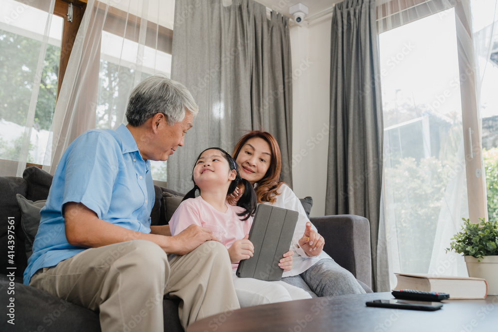 Asian grandparents and granddaughter using tablet at home. Senior Chinese, grandpa and grandma happy spend family time relax with young girl checking social media, lying on sofa in living room concept Asian grandparents and granddaughter using tablet at home. Senior Chinese, grandpa and grandma happy spend family time relax with young girl checking social media, lying on sofa in living room concept