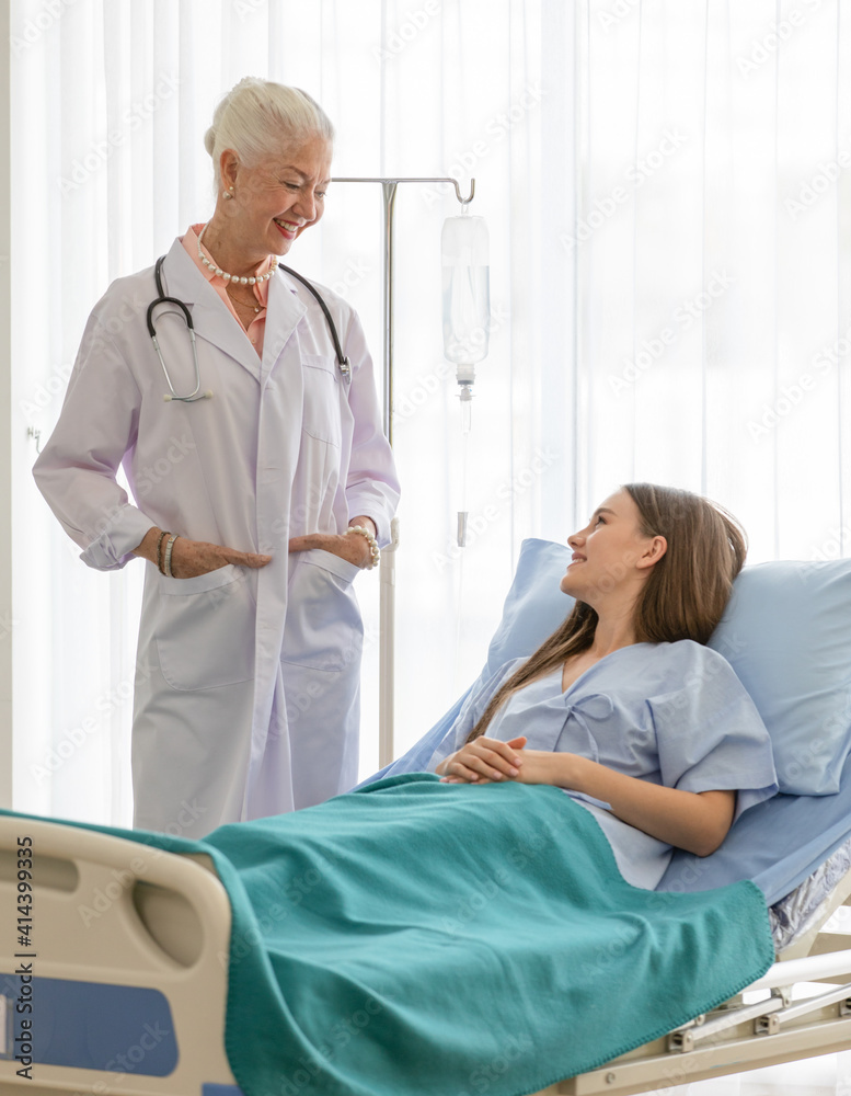 A senior older female doctor standing in an elegant pose put hands in pocket, and smile with kindness taking photos with a younger patient. A senior older female doctor standing in an elegant pose put hands in pocket, and smile with kindness taking photos with a younger patient.