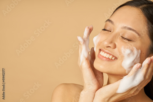Beauty portrait of lovely young woman smiling with eyes closed while applying gentle foam facial cleanser isolated over beige background