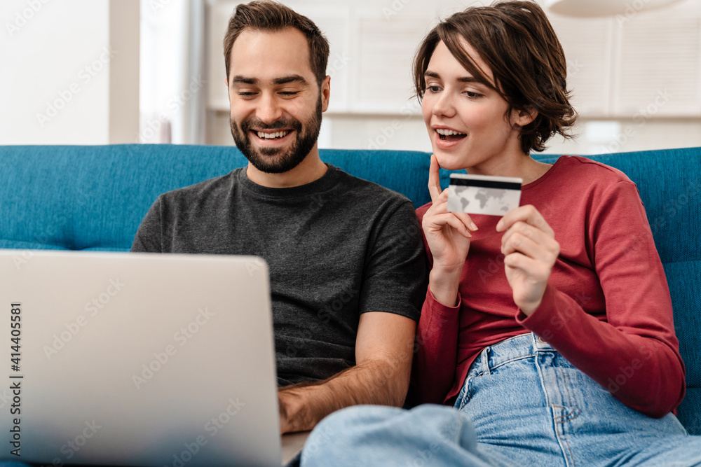 Happy young couple using laptop and credit card while sitting on couch Happy young couple using laptop and credit card while sitting on couch