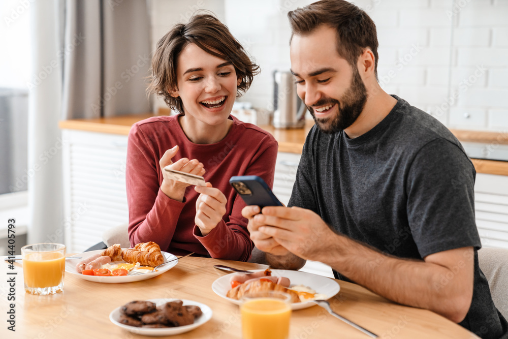 Joyful beautiful couple using cellphone and credit card while having breakfast Joyful beautiful couple using cellphone and credit card while having breakfast