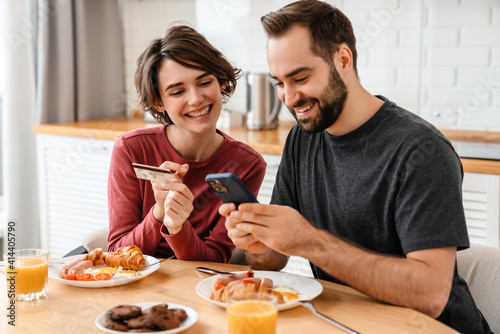 Joyful beautiful couple using cellphone and credit card while having breakfast