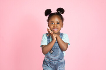 Happy laughing black skinned little girl in casual wear, jeans overalls, looking at camera, holding hands together. Little child with afrohair on pink studio background
