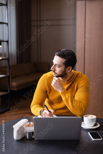 pensive arabian freelancer holding pen near laptop and looking away in restaurant