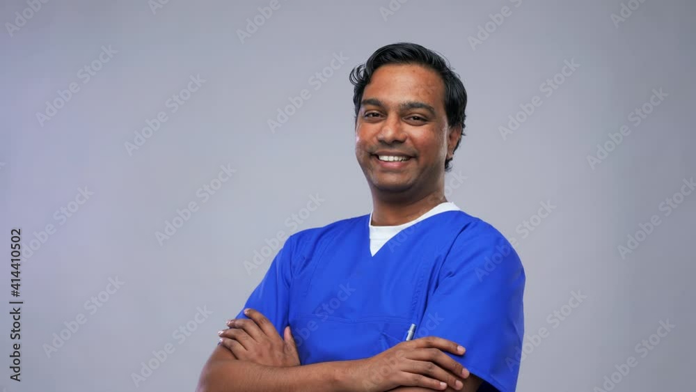 healthcare, profession and medicine concept - happy smiling indian doctor or male nurse in blue uniform with crossed arms over grey background healthcare, profession and medicine concept - happy smiling indian doctor or male nurse in blue uniform with crossed arms over grey background