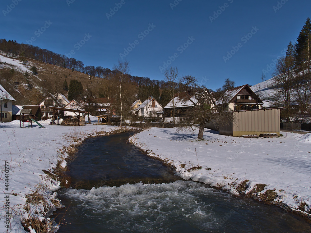 Idyllic winter landscape of small rural village Gundelfingen, part of Münsingen, Germany in Swabian Alb with Große Lauter river, snow-covered meadow and residential houses on sunny day with blue sky. Idyllic winter landscape of small rural village Gundelfingen, part of Münsingen, Germany in Swabian Alb with Große Lauter river, snow-covered meadow and residential houses on sunny day with blue sky.