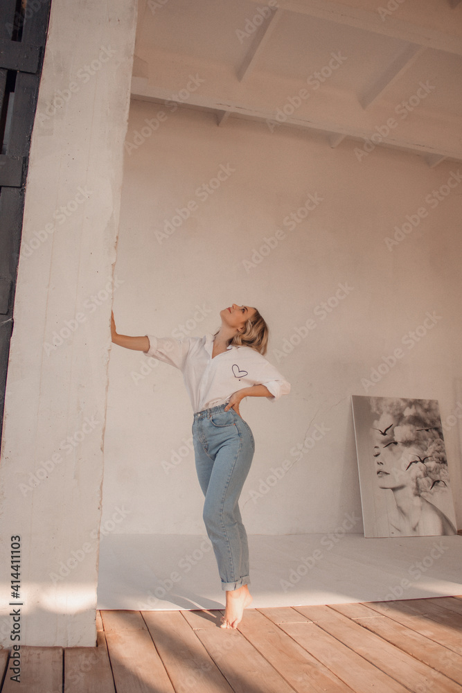 young glamour blonde woman with long legs in jeans and white shirt is standing in profile on the wooden floor and looking away with smile on white wall background, lifestyle concept, free space young glamour blonde woman with long legs in jeans and white shirt is standing in profile on the wooden floor and looking away with smile on white wall background, lifestyle concept, free space