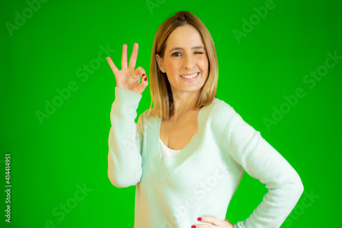 Young beautiful woman wearing casual sweater standing over green isolated background smiling positive doing okay sign with hand and fingers. Successful expression.
