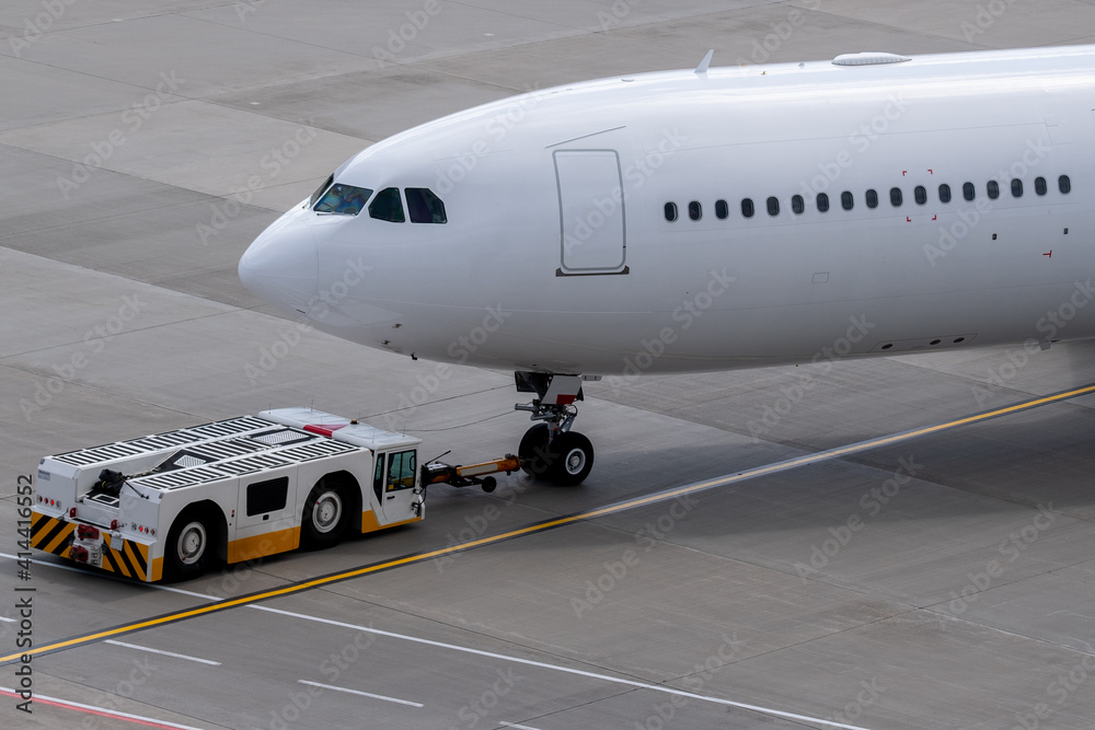 The apron transporter tows a modern narrow-body white passenger aircraft across the airfield of an international airport.