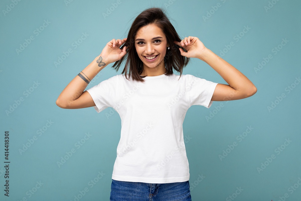 Portrait of positive young beautiful brunette woman with sincere emotions wearing casual white t-shirt for mockup isolated over blue background with copy space - obrazy, fototapety, plakaty 