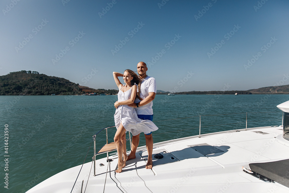 Amazing girl in a white summer dress and an handsome man posing on the corner of their white yacht. Man is hugging his lovely wife on his yacht against the backdrop of water and nature.