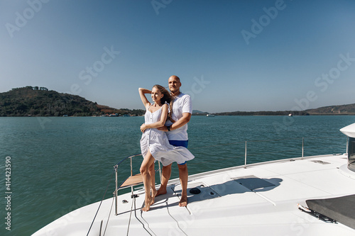 Amazing girl in a white summer dress and an handsome man posing on the corner of their white yacht. Man is hugging his lovely wife on his yacht against the backdrop of water and nature. © Semachkovsky 