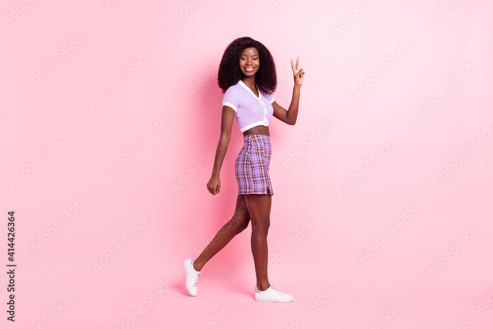 Full length body size profile side view of pretty cheerful thin girl walking showing v-sign isolated over pink pastel color background Full length body size profile side view of pretty cheerful thin girl walking showing v-sign isolated over pink pastel color background