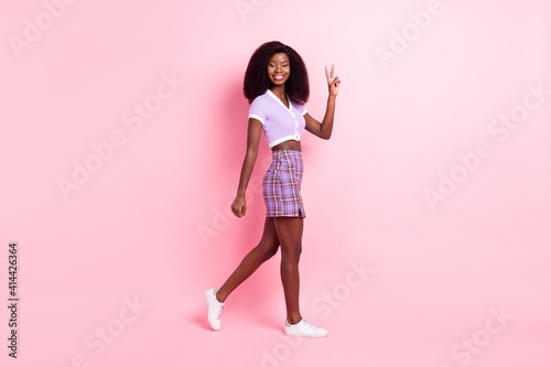 Full length body size profile side view of pretty cheerful thin girl walking showing v-sign isolated over pink pastel color background © deagreez