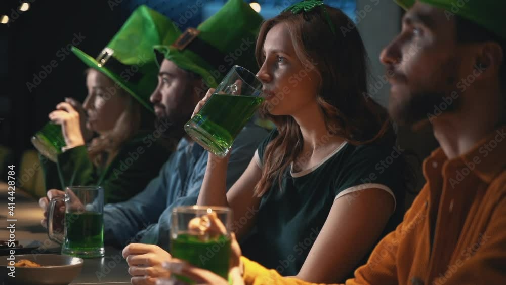 Friends watching TV in a pub and drinking beer. Men and women having fun in Irish hats. Saint Patrick's Day.