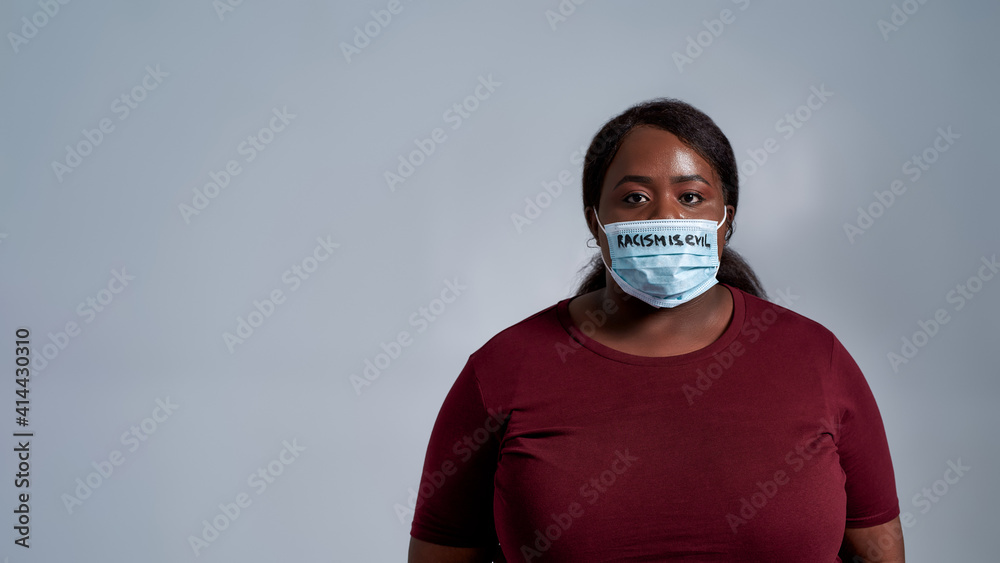 Serious plus size young african american woman in casual clothes wearing protective face mask with Racism is evil text looking at camera, posing isolated over gray background Serious plus size young african american woman in casual clothes wearing protective face mask with Racism is evil text looking at camera, posing isolated over gray background