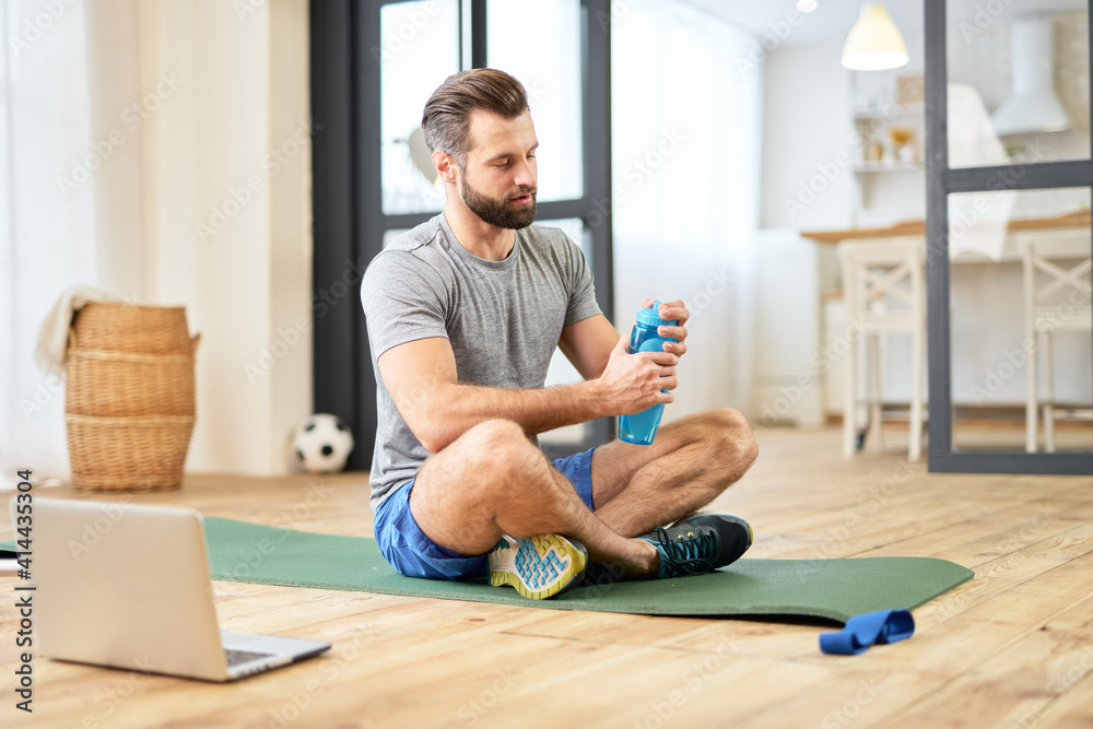 Athletic young man with bottle of water sitting in exercise mat