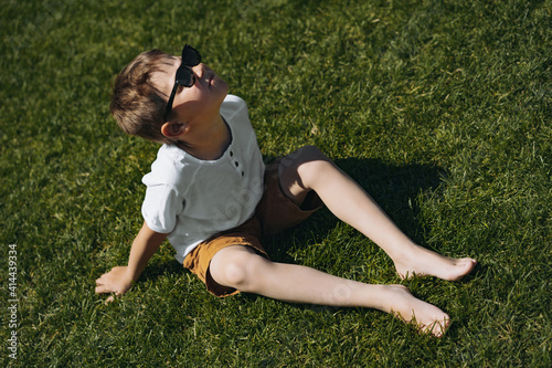 cute caucasian boy wearing white henley tee shirt and sunglasses sitting on green grass and looking up into the sky. photo