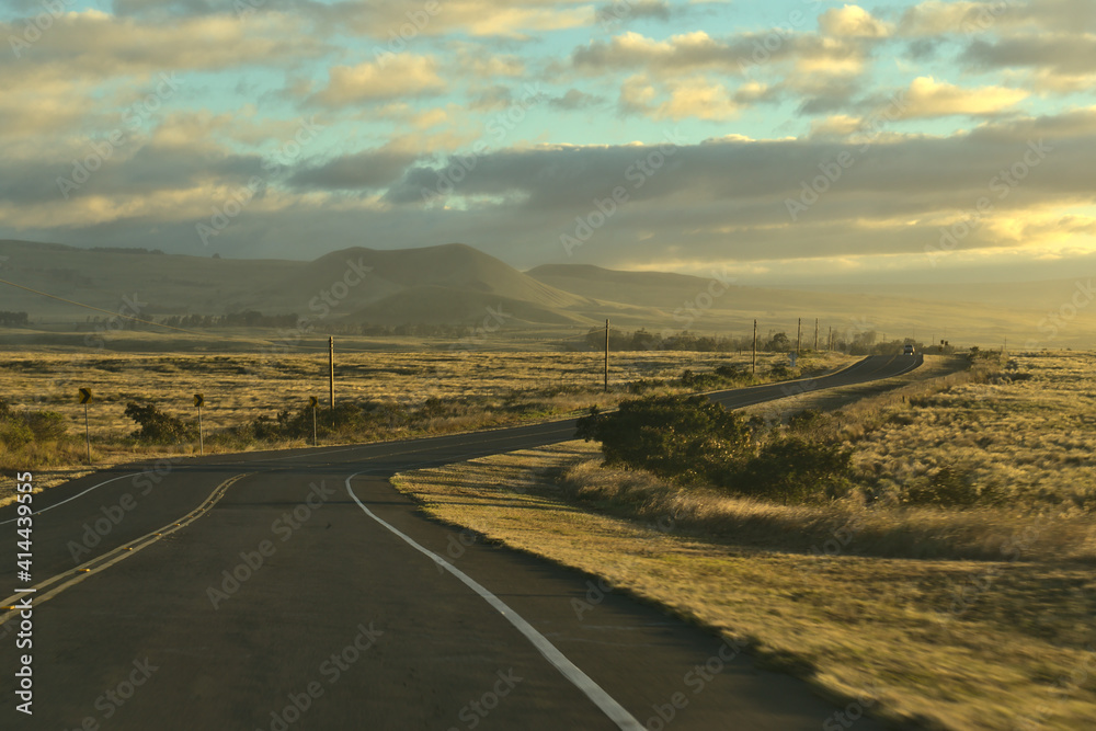 Driving on a scenic, windy road on the north western part of Big Island, Hawai'i at sunset, with golden fields, hazy mountains and cloudy gray blue sky in the background