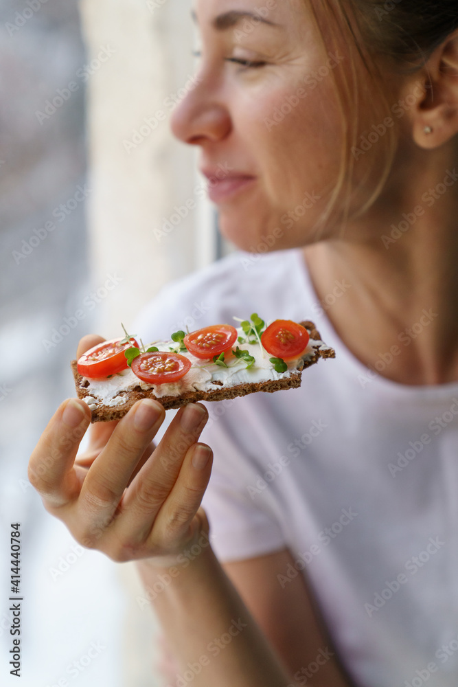 Happy woman eating rye crisp bread with creamy vegetarian cheese tofu, cherry tomato and rucola micro greens, sitting at home and looking at window. Healthy food, gluten free, diet concept. Happy woman eating rye crisp bread with creamy vegetarian cheese tofu, cherry tomato and rucola micro greens, sitting at home and looking at window. Healthy food, gluten free, diet concept.