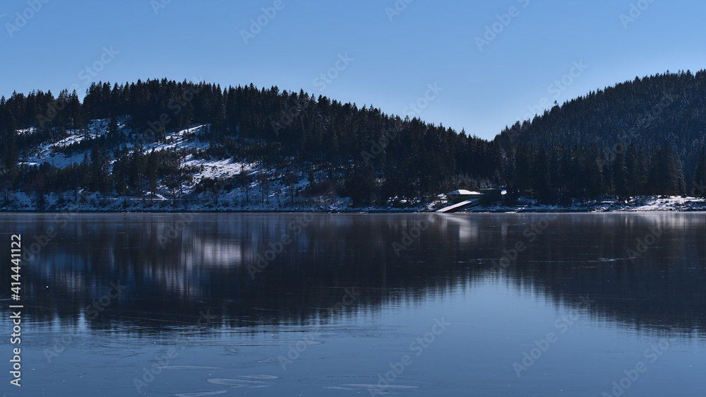Peaceful view of frozen lake Schluchsee in the Black Forest hills, Germany in winter season with coniferous forest reflected on the ice surface on sunny day with blue sky.