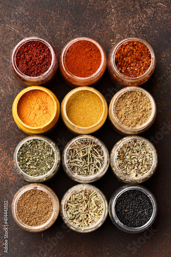 Fototapeta Naklejka Na Ścianę i Meble -  Set of various multicolored spices in jars over dark brown concrete background. Top view, close up, flat lay. Food background, ingredients for cooking, assorted condiment