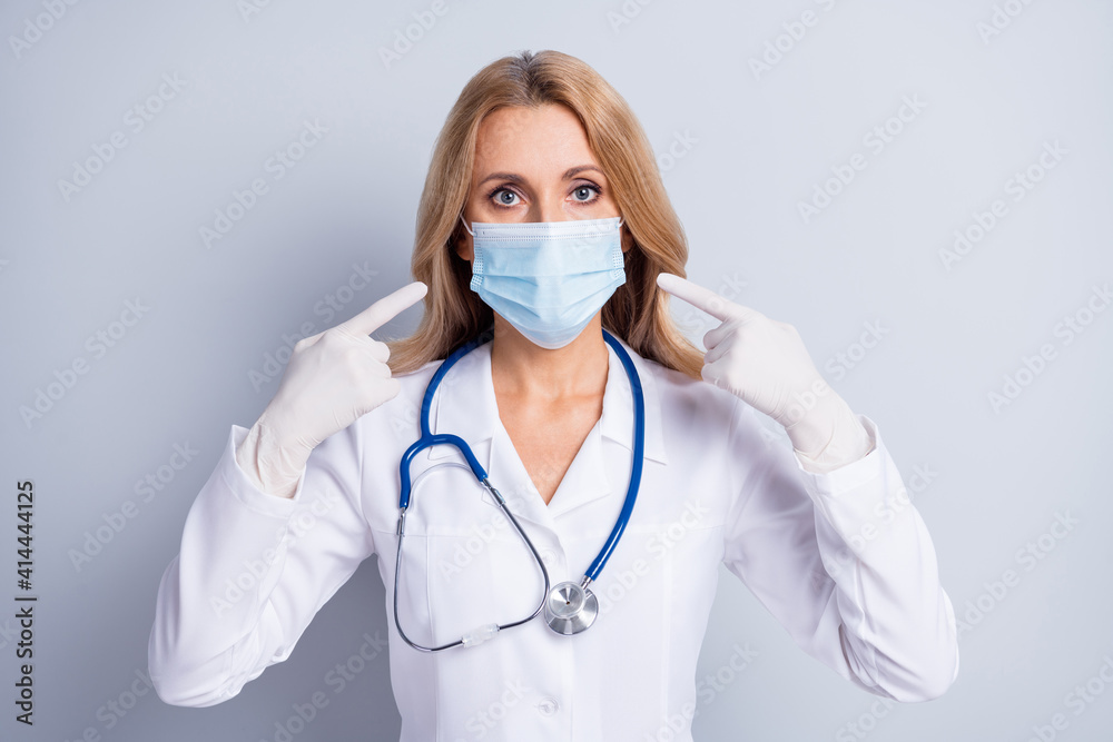 Photo of mature woman doctor wear rubber gloves point finger at face mask coronavirus isolated on grey color background Photo of mature woman doctor wear rubber gloves point finger at face mask coronavirus isolated on grey color background