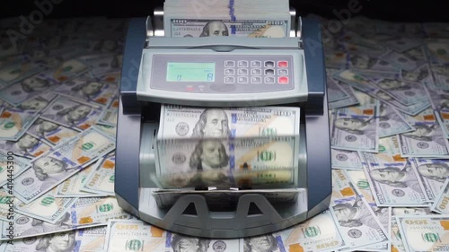 Currency counting machine stands on heap of hundred dollars. Cash bill counter money machine counts 100 dollar bills or USD banknotes. Automatic mechanism for bank financial operations. Slow motion