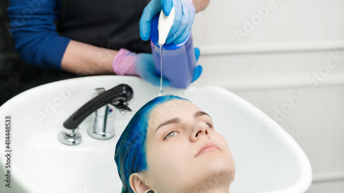 Fototapeta Naklejka Na Ścianę i Meble -  Hairstylist hands in gloves squeezes shampoo from tube into head young woman with blue hair while washing hair in special sink and shower at beauty salon after hair dyeing process.