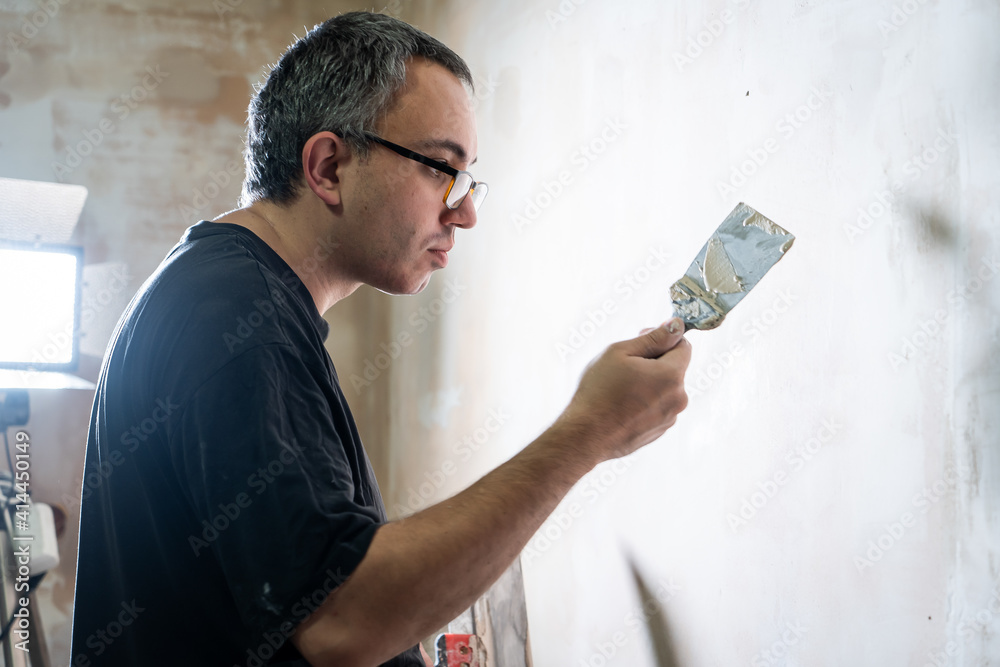 Manual worker with wall plastering tools renovating house. Plasterer renovating indoor walls and ceilings with float and plaster. Happy worker man making wall in the room by hand spatula Manual worker with wall plastering tools renovating house. Plasterer renovating indoor walls and ceilings with float and plaster. Happy worker man making wall in the room by hand spatula