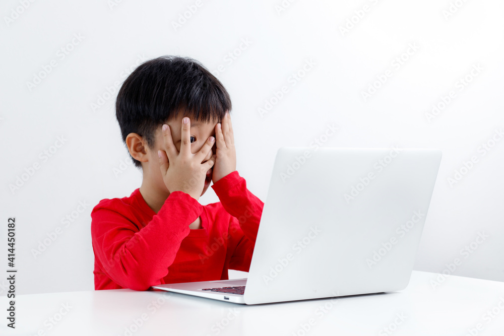 Portrait of a surprised little Asian boy peeking while watching something on a laptop, sitting at a desk, isolated on white background. Portrait of a surprised little Asian boy peeking while watching something on a laptop, sitting at a desk, isolated on white background.