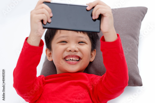 Happy little Asian boy lying on a pillow, smiling and using a smartphone at home, isolated on white background.