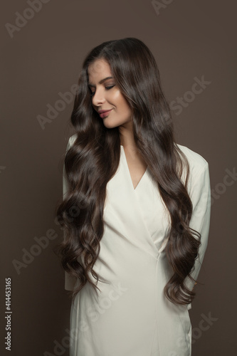 Nice woman with long dark hairstyle wearing white dress standing on brown background