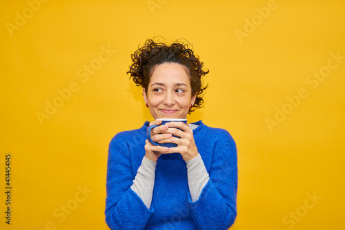 Happy girl drinking hot beverage. Pretty young woman enjoying drink. Holding blue cap with both hands. Wearing blue sweater over isolated yellow background. Curly hair.