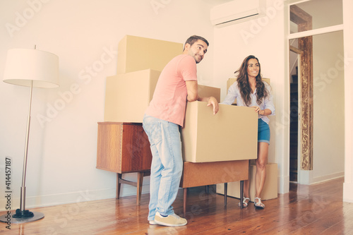 Positive young couple looking over their new apartment, while standing and leaning on cardboard boxes and furniture indoors. Full length. New home or property buying concept © Mangostar