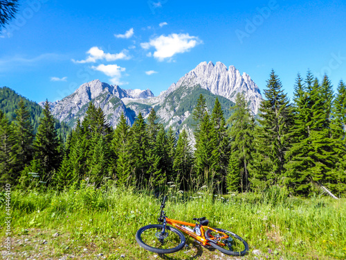 High Alpine mountain biking. A bike is lying on the ground, next to a small gravelled pathway. In the back there are tall Lienz Dolomites. Pine trees disturbing the view on mountain range. Adrenaline