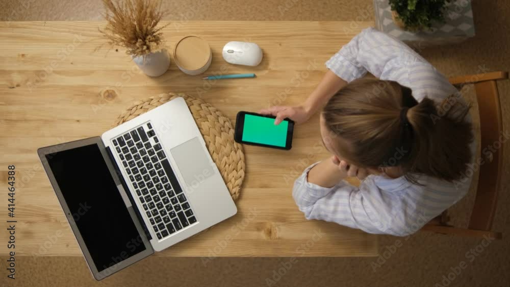 The girl looks at the phone with a green screen. Opposite is an open laptop with a green screen. The concept of remote work from top view.